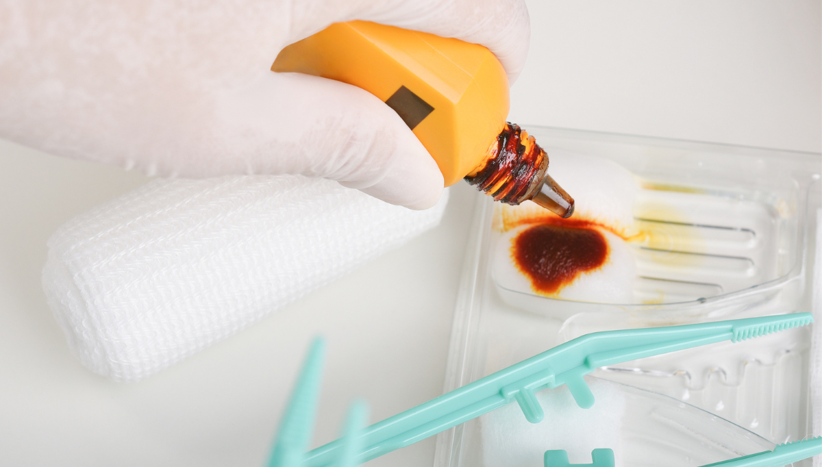 Mão a a garrar em frasco de iodopovidona em algodão, ligadura e pinça de plástico esverdeada.